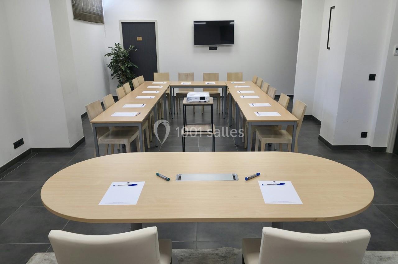 Salle de réunion avec tables disposées en U, chaises, écran mural, et documents avec stylos sur les tables.
