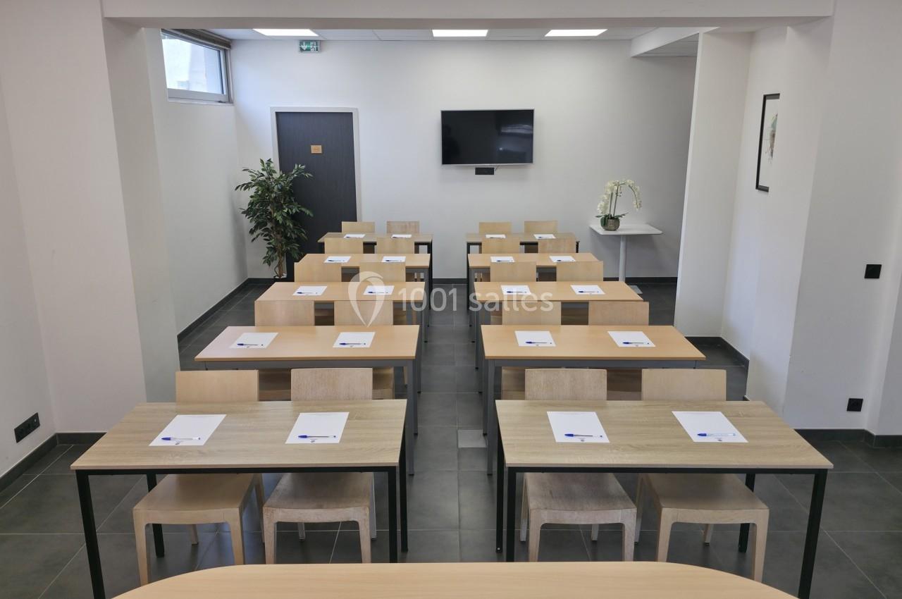 Salle de formation avec des tables en bois, des chaises alignées, un écran mural et des documents posés sur chaque table.