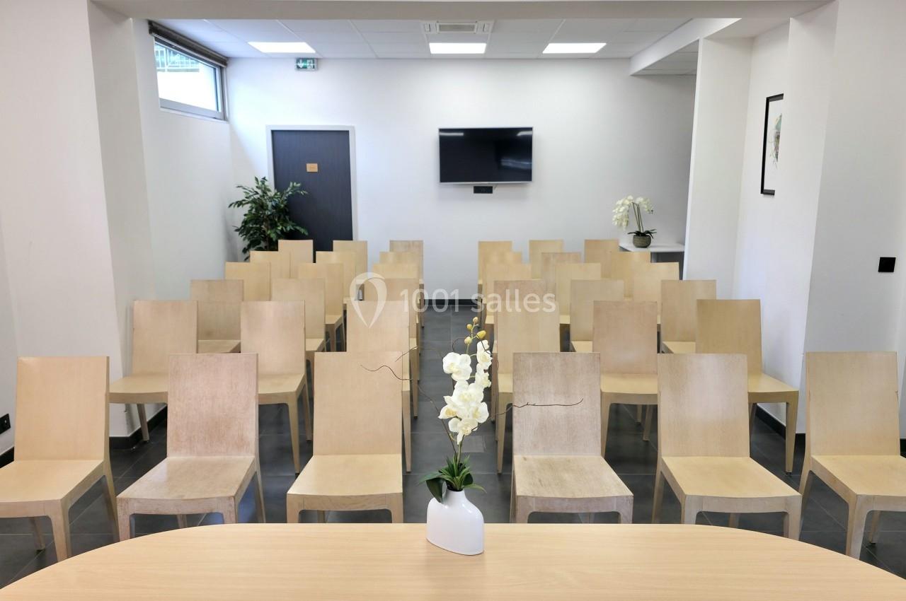 Salle de réunion avec des chaises en bois alignées, un écran mural et une table avec une orchidée au premier plan.