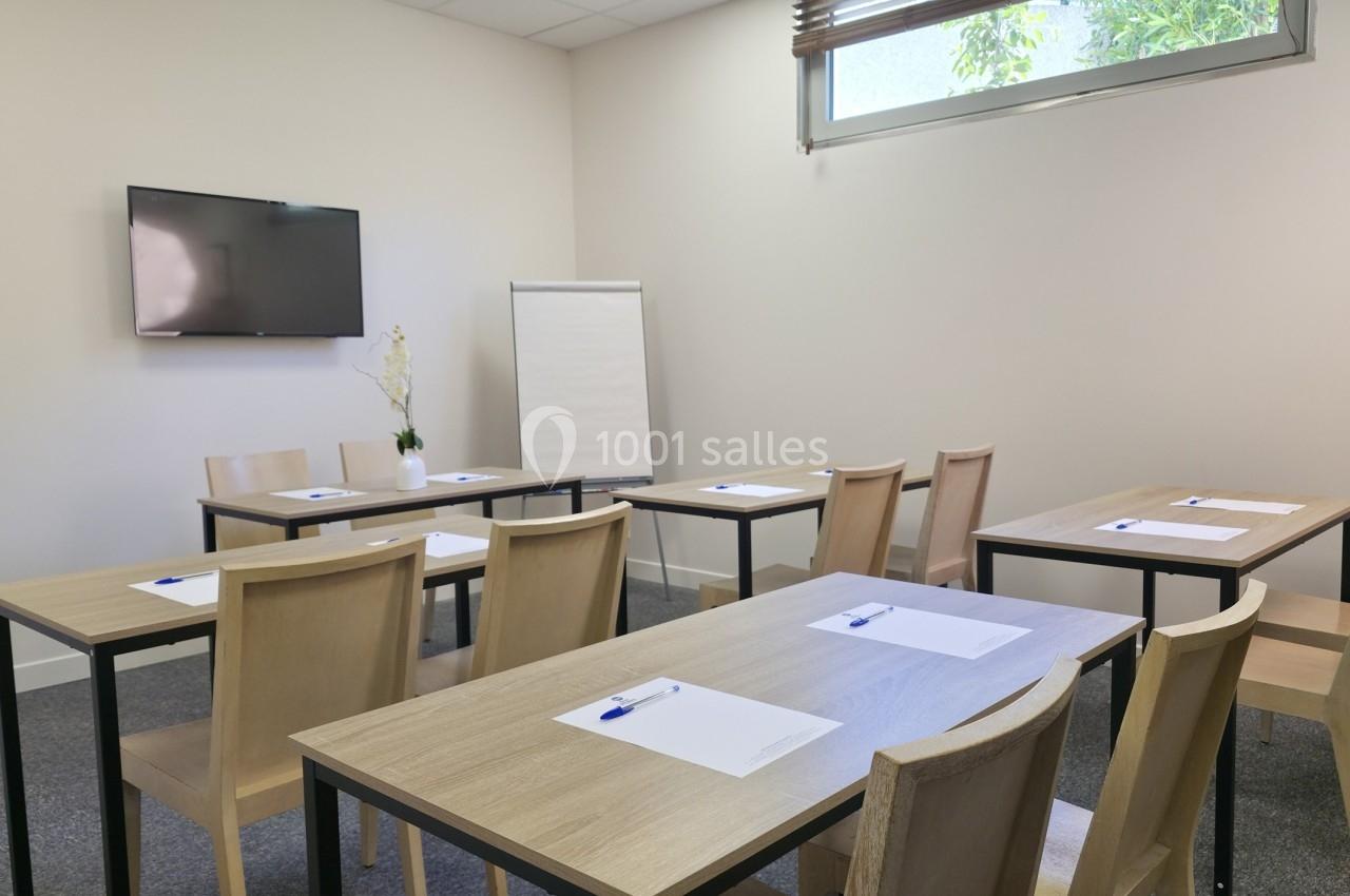 Salle de réunion lumineuse avec tables, chaises, tableau blanc, écran mural et fenêtre donnant sur l'extérieur.