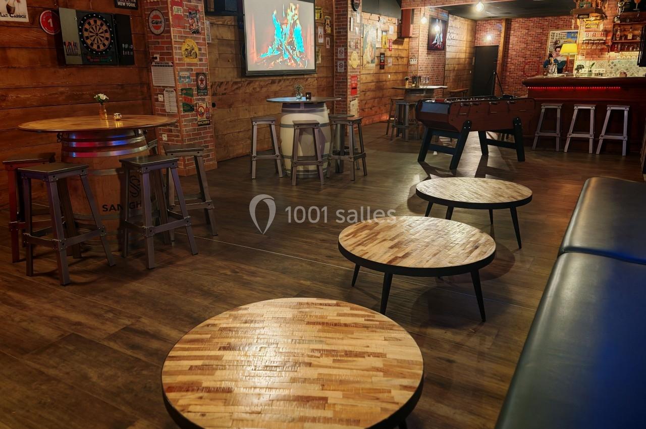 Salle de bar avec tables en bois, baby-foot, écran mural et décoration rétro sur des murs en briques et bois.