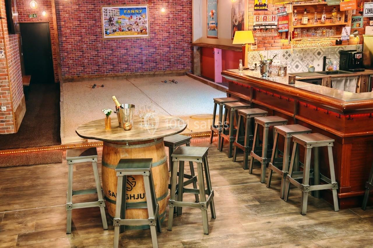 Salle de bar avec comptoir en bois, tabourets métalliques et espace de jeu de pétanque sur sol en sable.