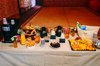 Table garnie de verrines, brochettes de fruits frais et plateaux de donuts colorés sur une nappe décorative.