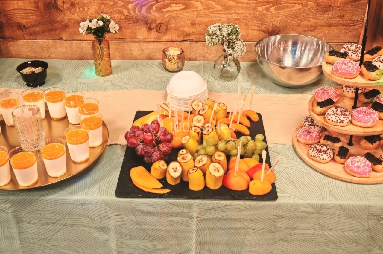 Table garnie de verrines, brochettes de fruits frais et plateaux de donuts colorés sur une nappe décorative.