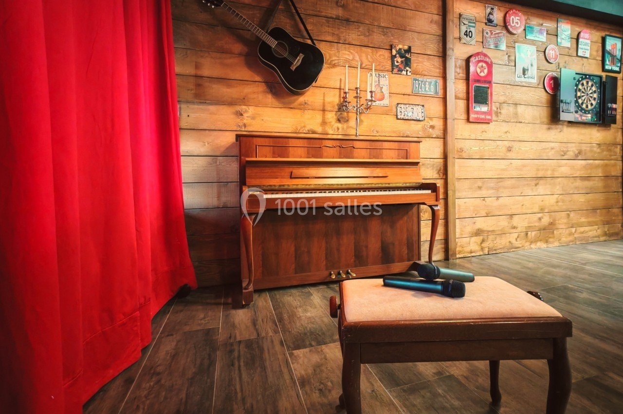 Un piano droit en bois devant un mur décoré, avec une guitare suspendue, un tabouret et un rideau rouge à gauche.