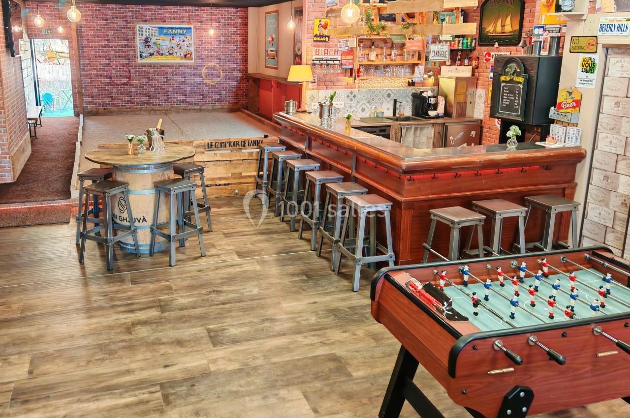 Salle de loisirs avec baby-foot, bar en bois, tabourets, terrain de pétanque intérieur et décoration rétro.