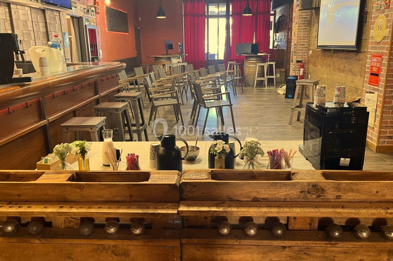 Salle aménagée avec des chaises disposées face à un écran, un bar en bois au premier plan avec des boissons et des…