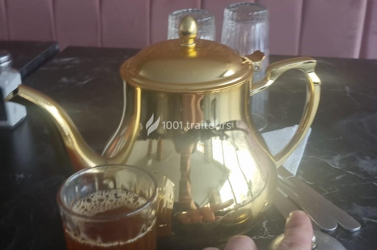 Théière dorée posée sur une table en marbre noir avec un verre de thé et des couverts à proximité.