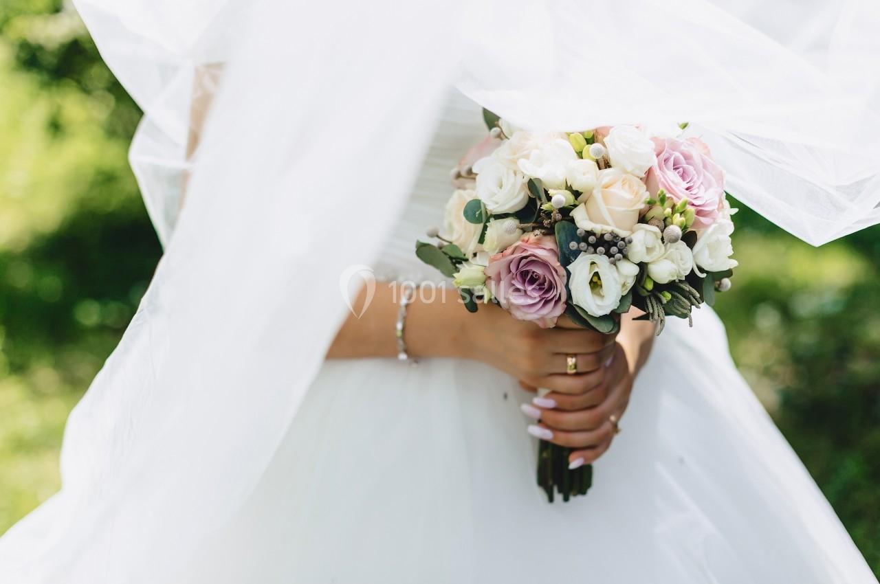 Une mariée en robe blanche tient un bouquet de fleurs pastel, avec un voile flottant en arrière-plan.
