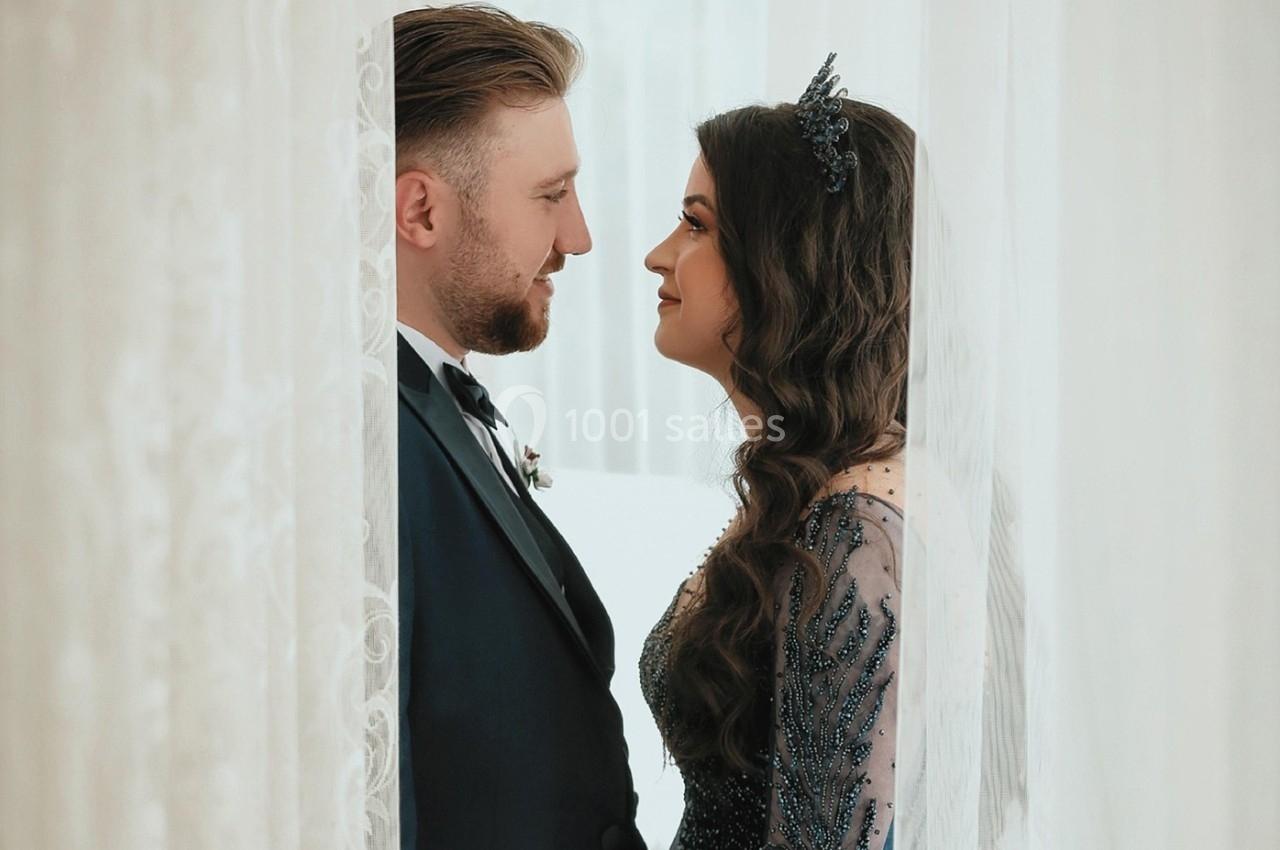 Un couple habillé élégamment se regarde avec tendresse entre deux rideaux blancs.