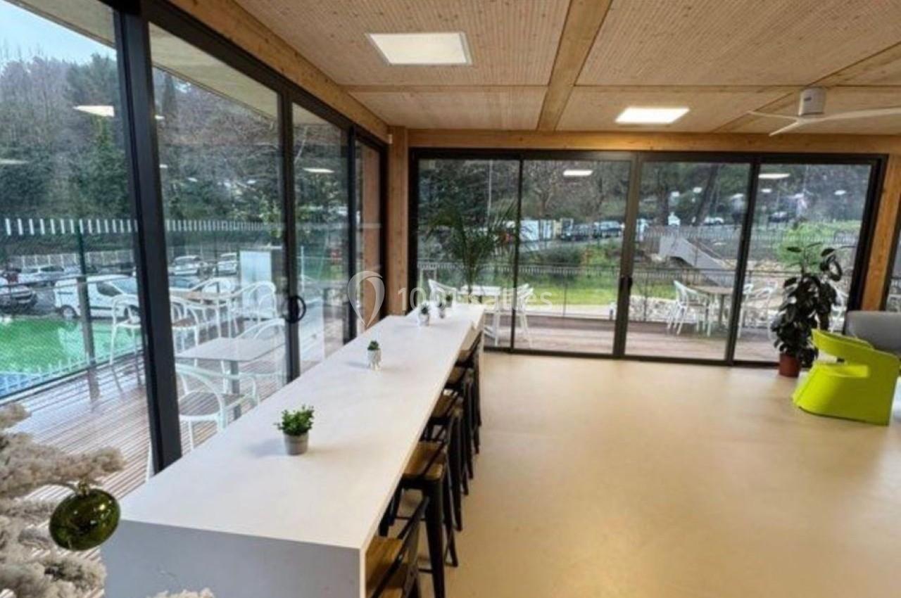 Salle lumineuse avec de grandes baies vitrées, une table haute entourée de tabourets et vue sur un espace extérieur…