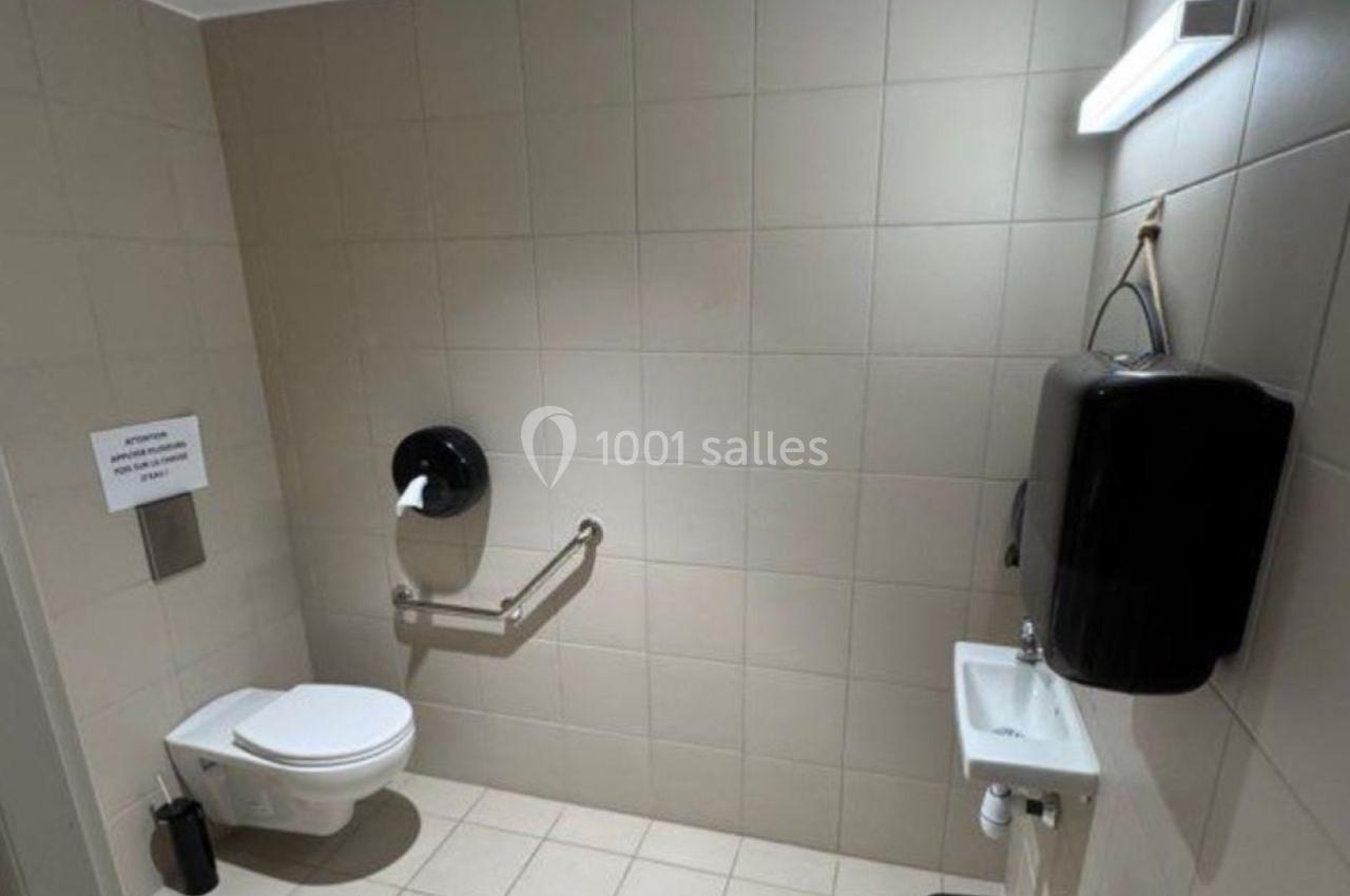 Toilettes accessibles avec barre d'appui, lavabo, distributeur de savon et poubelle sur un sol carrelé beige.