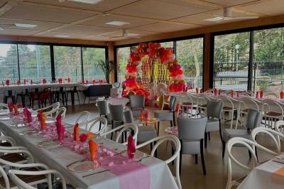 Salle décorée avec des guirlandes colorées et des lanternes, tables dressées pour un événement festif près de grandes baies…