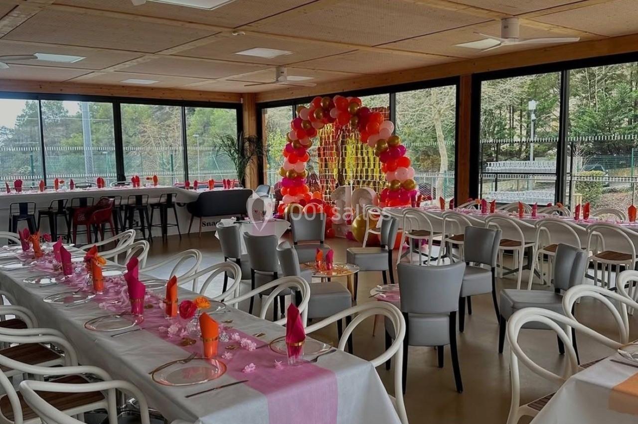 Salle décorée pour un événement avec tables dressées, serviettes colorées et arche de ballons en arrière-plan.