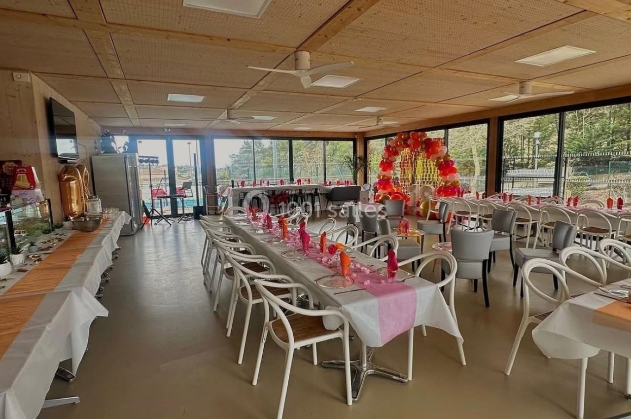 Salle décorée pour un événement avec tables alignées, nappes blanches et roses, et arche florale en arrière-plan.