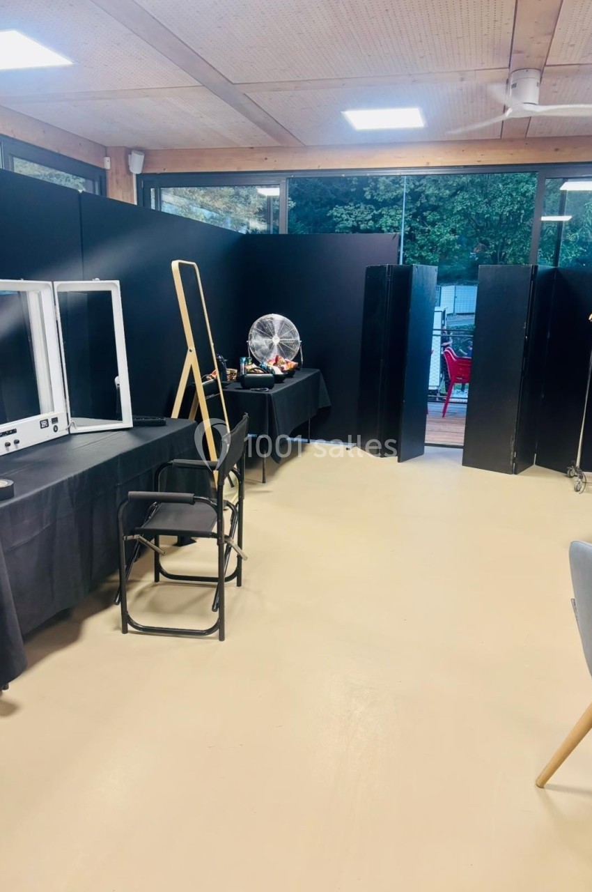 Salle aménagée avec des tables, chaises, panneaux noirs, ventilateur et matériel divers, éclairée par des baies vitrées.