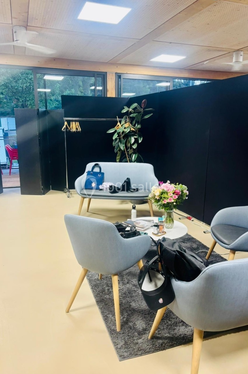 Espace salon avec fauteuils gris, table basse, fleurs, sacs et cintres en arrière-plan dans une pièce lumineuse.