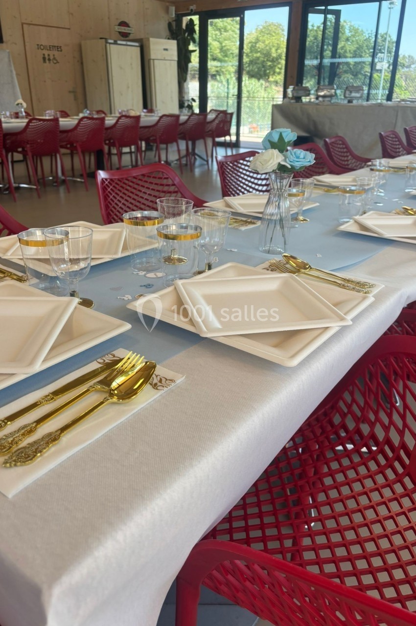 Table dressée avec vaisselle blanche, couverts dorés et décoration florale, dans une salle lumineuse avec chaises rouges.
