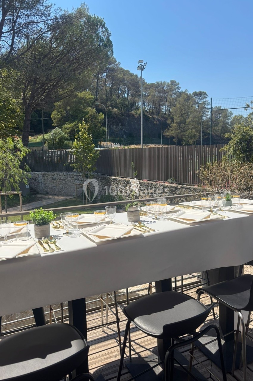 Table dressée en extérieur avec nappes blanches, verres et couverts, entourée de végétation et d'une clôture en bois.