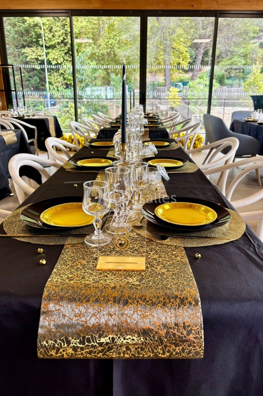 Table dressée avec nappes noires, chemins dorés, assiettes jaunes et verres, dans une salle lumineuse avec vue sur jardin.