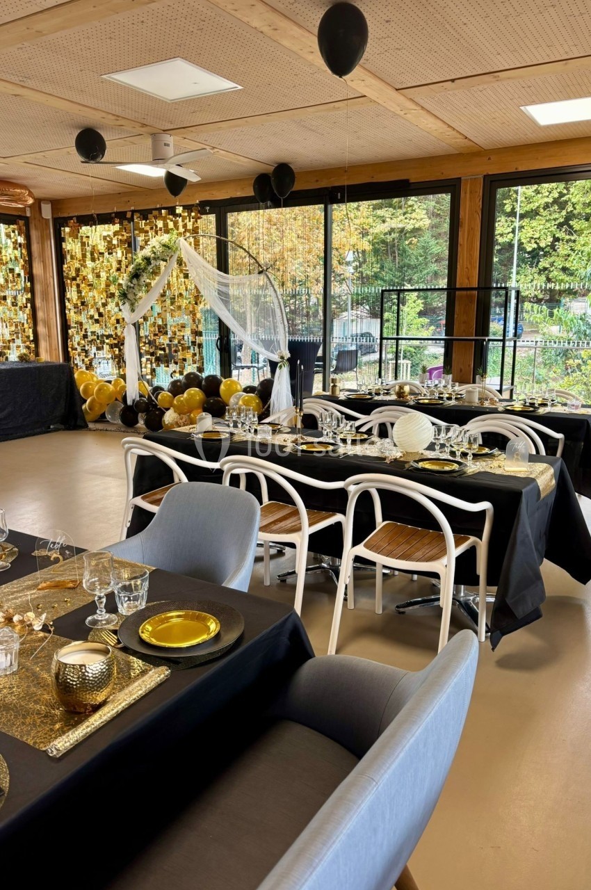 Salle décorée pour un événement avec tables dressées en noir et or, ballons suspendus et arche blanche en arrière-plan.