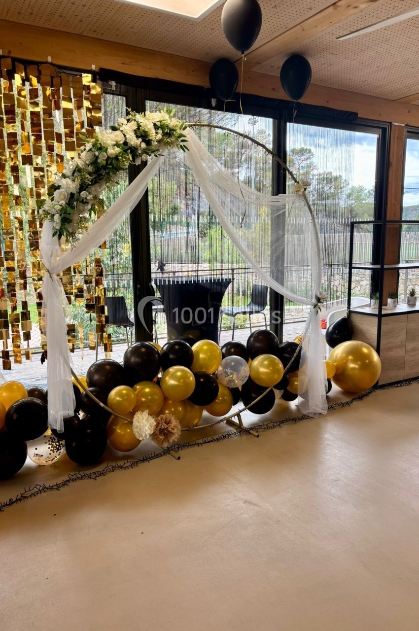 Arche décorative ornée de fleurs blanches, de voilage et de ballons noirs et dorés dans une salle lumineuse.