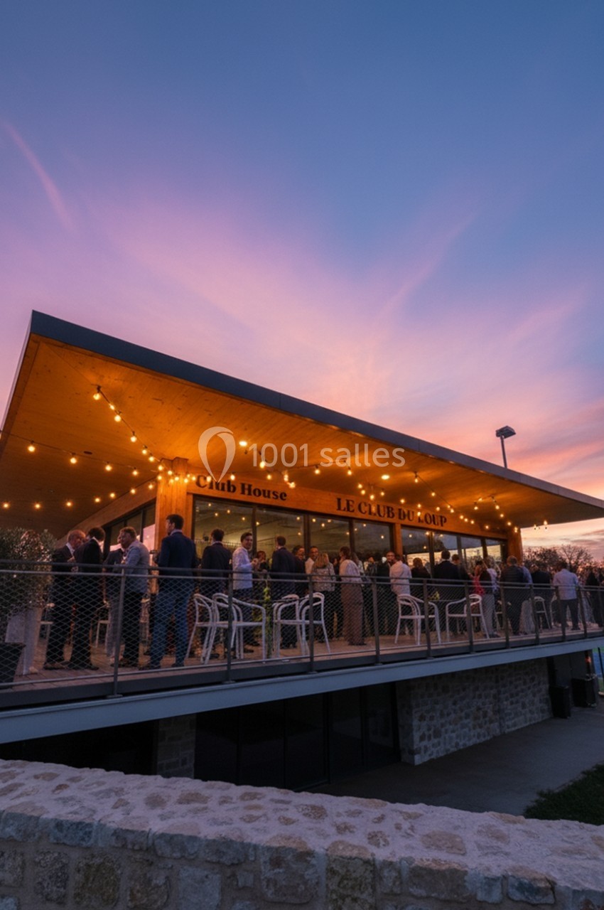 Terrasse d'un club-house moderne éclairée au crépuscule, avec des personnes rassemblées sous un ciel coloré.