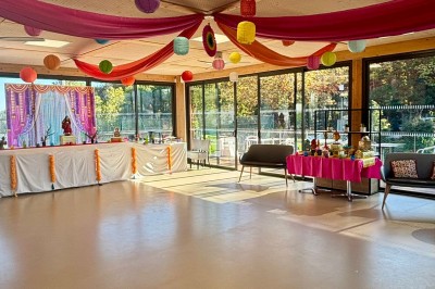 Salle décorée avec des guirlandes colorées et des lanternes, tables dressées pour un événement festif près de grandes baies…