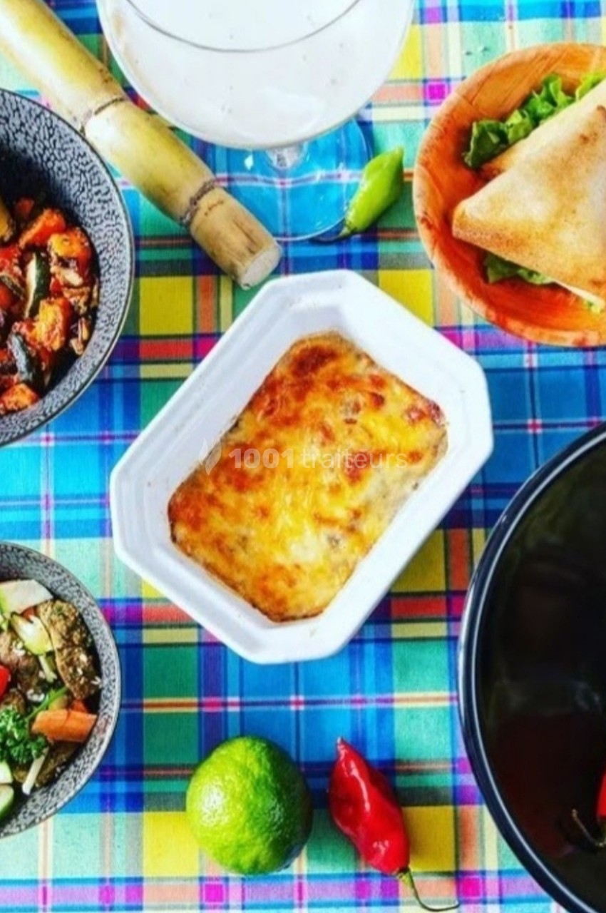 Repas créole coloré avec gratin, samoussas, légumes, brochettes et accompagnements sur une nappe à carreaux.
