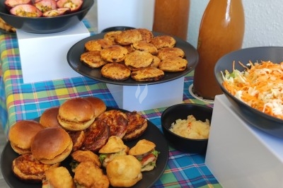 Buffet avec assiettes de mini-burgers, beignets, crudités, jus en bouteilles et salades sur une nappe colorée.