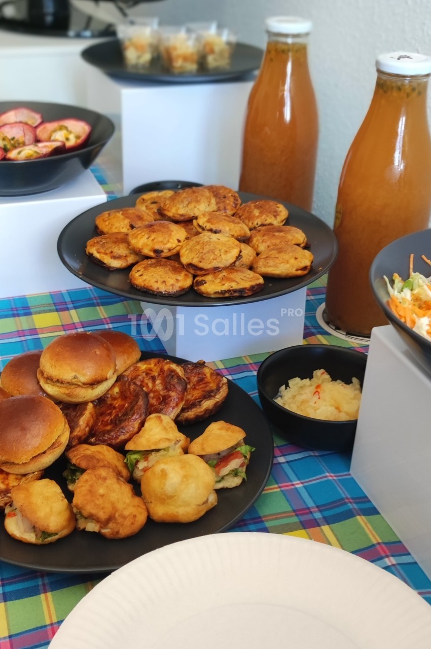 Buffet avec assiettes de mini-burgers, beignets, crudités, jus en bouteilles et salades sur une nappe colorée.