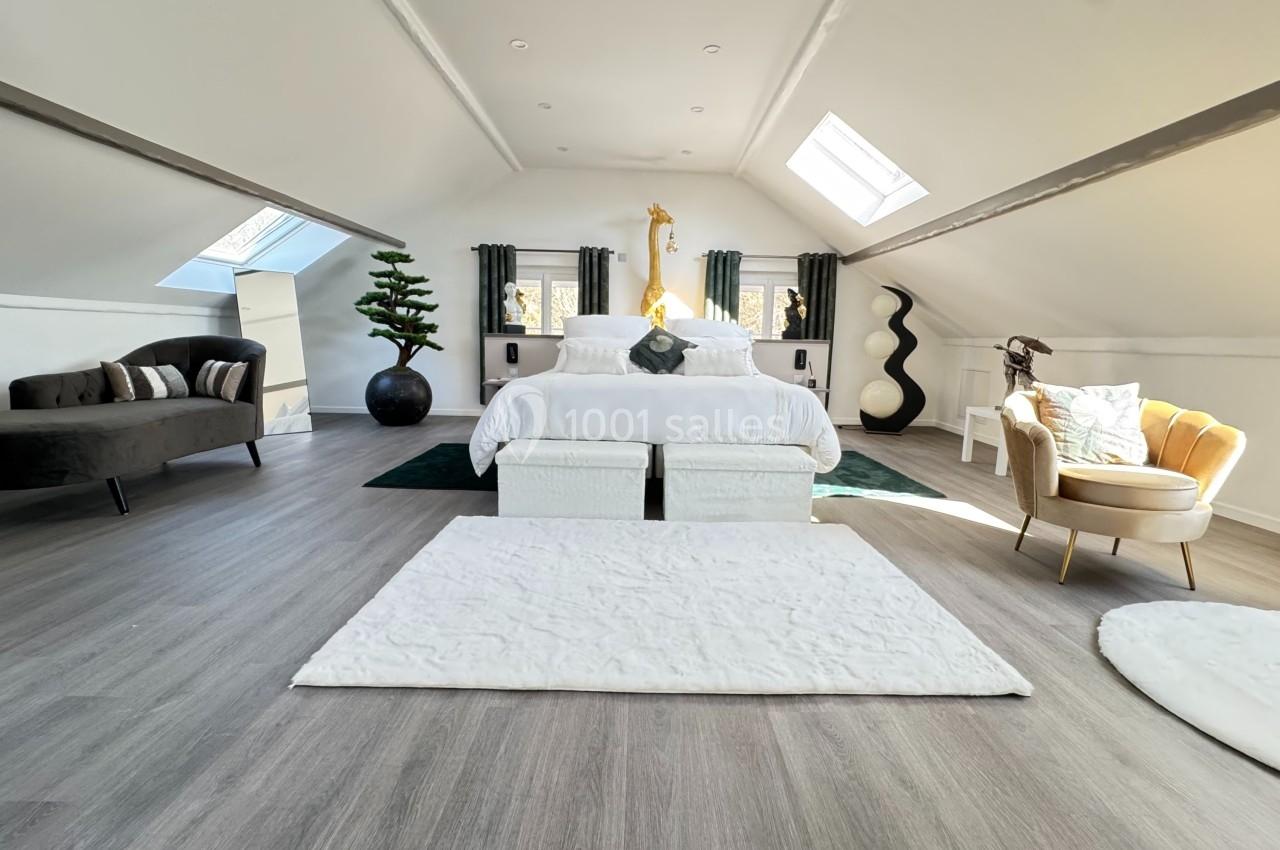 Chambre mansardée lumineuse avec lit central, fauteuils, canapé, décoration moderne et puits de lumière.