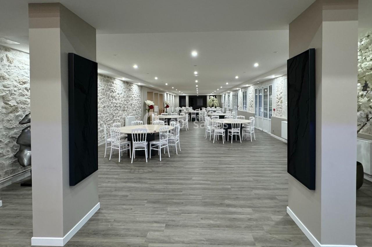 Salle de réception spacieuse avec murs en pierre, parquet et tables rondes dressées de chaises blanches.