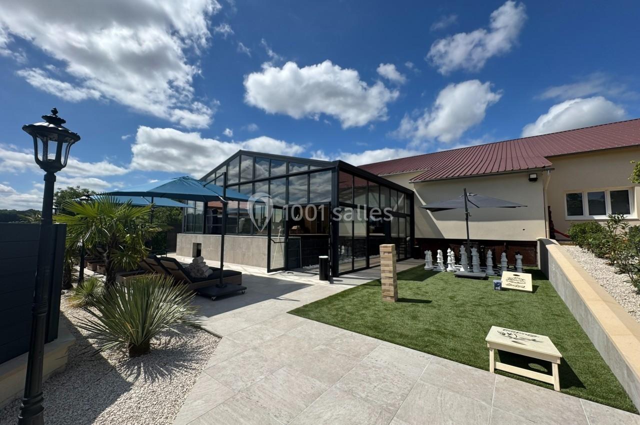 Cour extérieure avec terrasse, pelouse artificielle, échiquier géant, jeu de Jenga et mobilier sous un ciel ensoleillé.