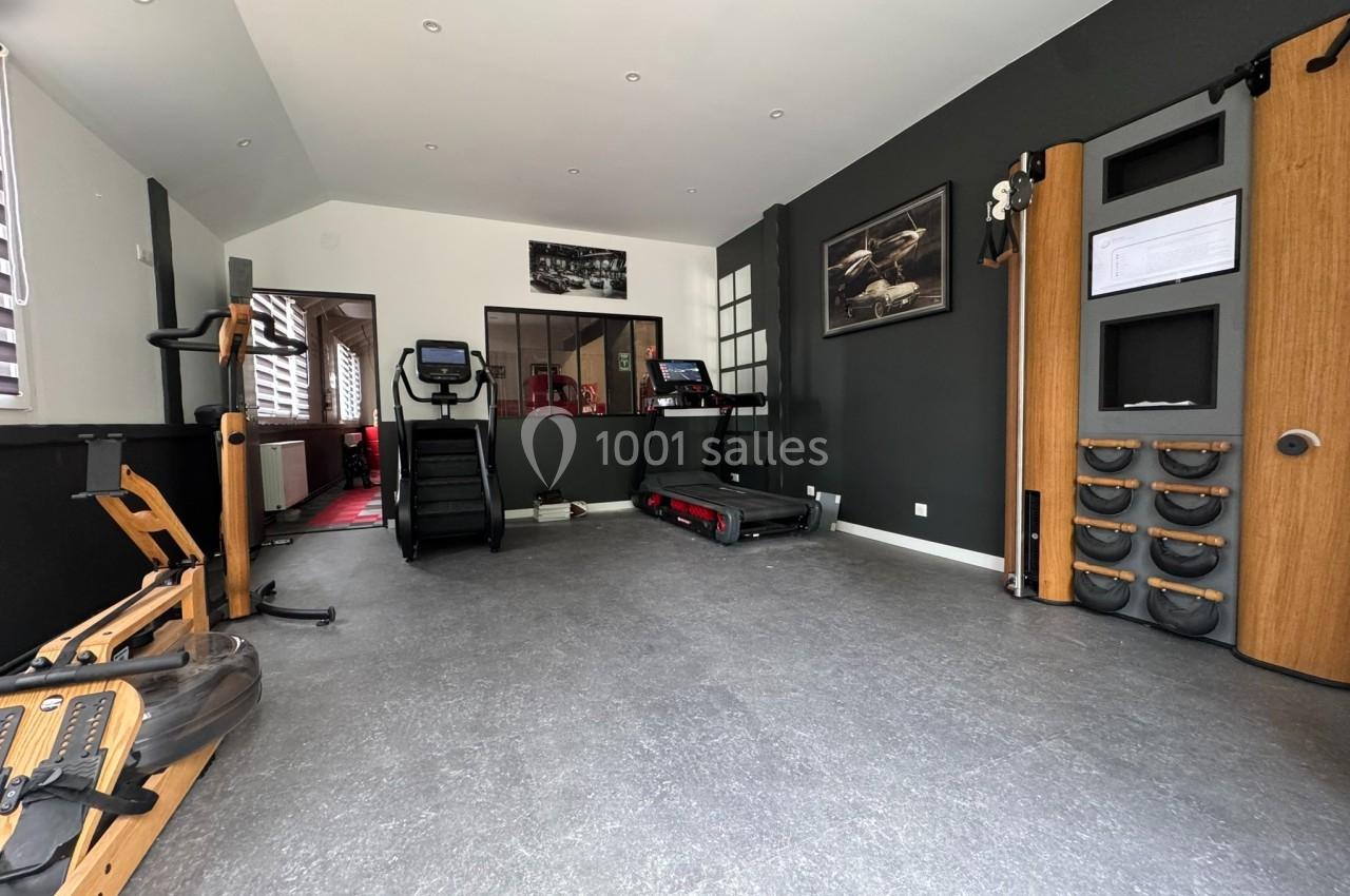 Salle de sport équipée de tapis de course, rameurs et matériel de musculation, avec murs sombres et éclairage naturel.