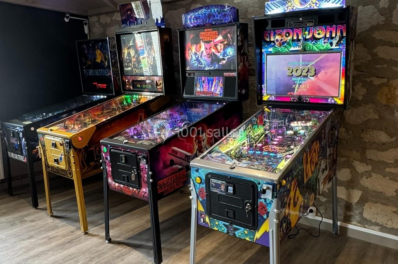 Quatre flippers alignés dans une salle au mur en pierre, éclairés par une lumière tamisée.