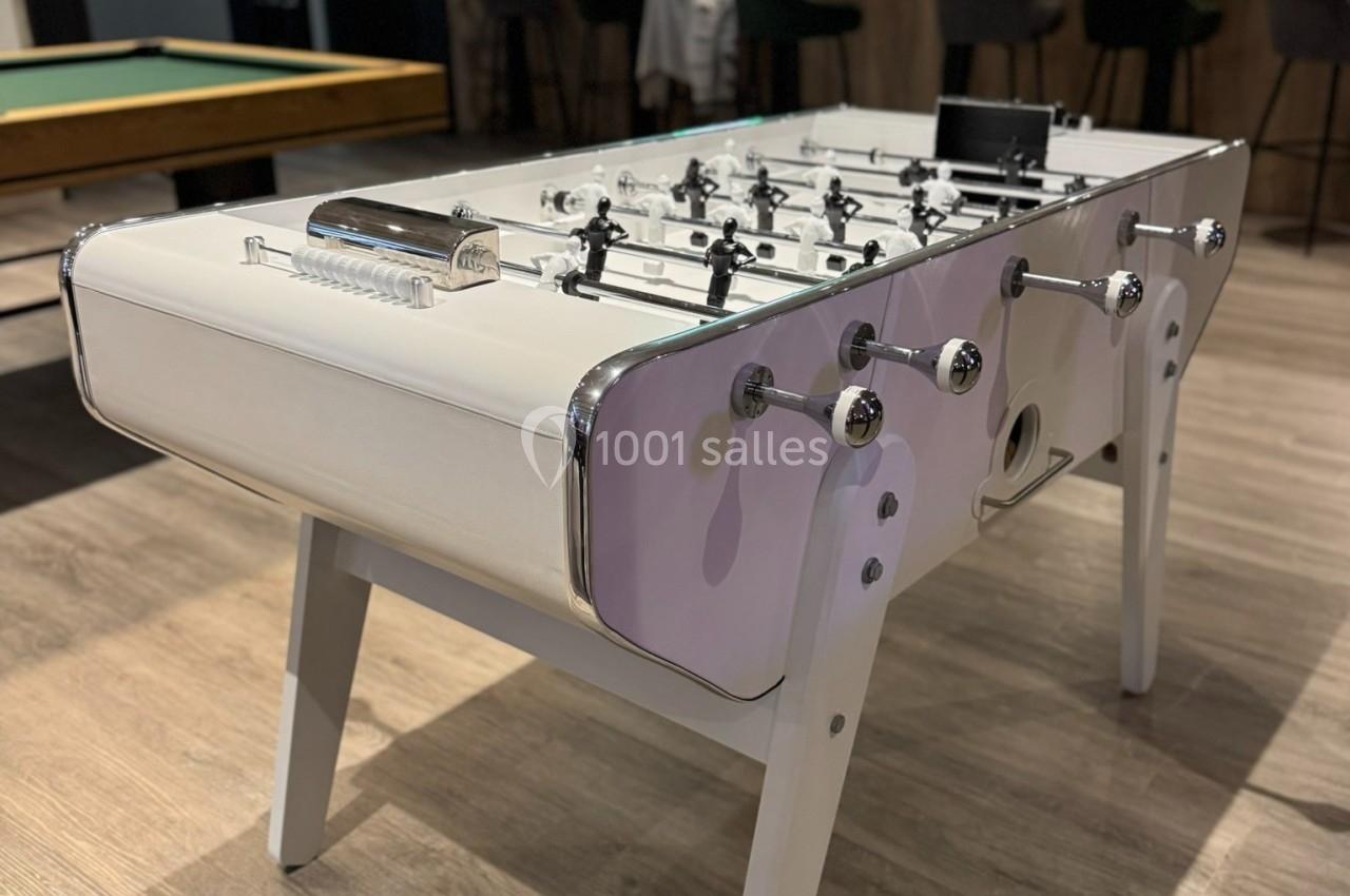 Table de baby-foot blanche dans une salle de loisirs avec un bar et une table de billard en arrière-plan.
