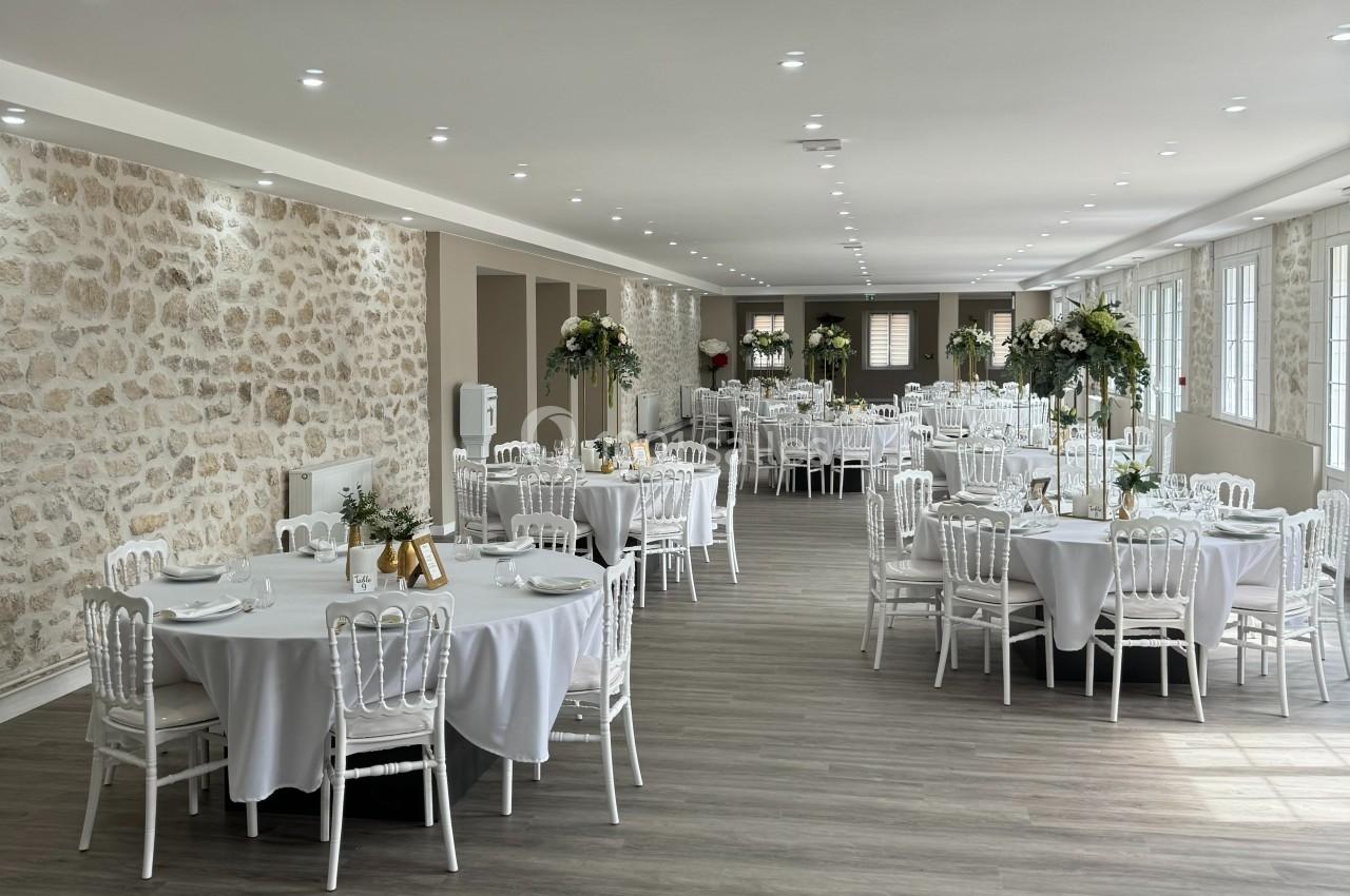 Salle de réception lumineuse avec tables rondes dressées, nappes blanches et décorations florales élégantes.