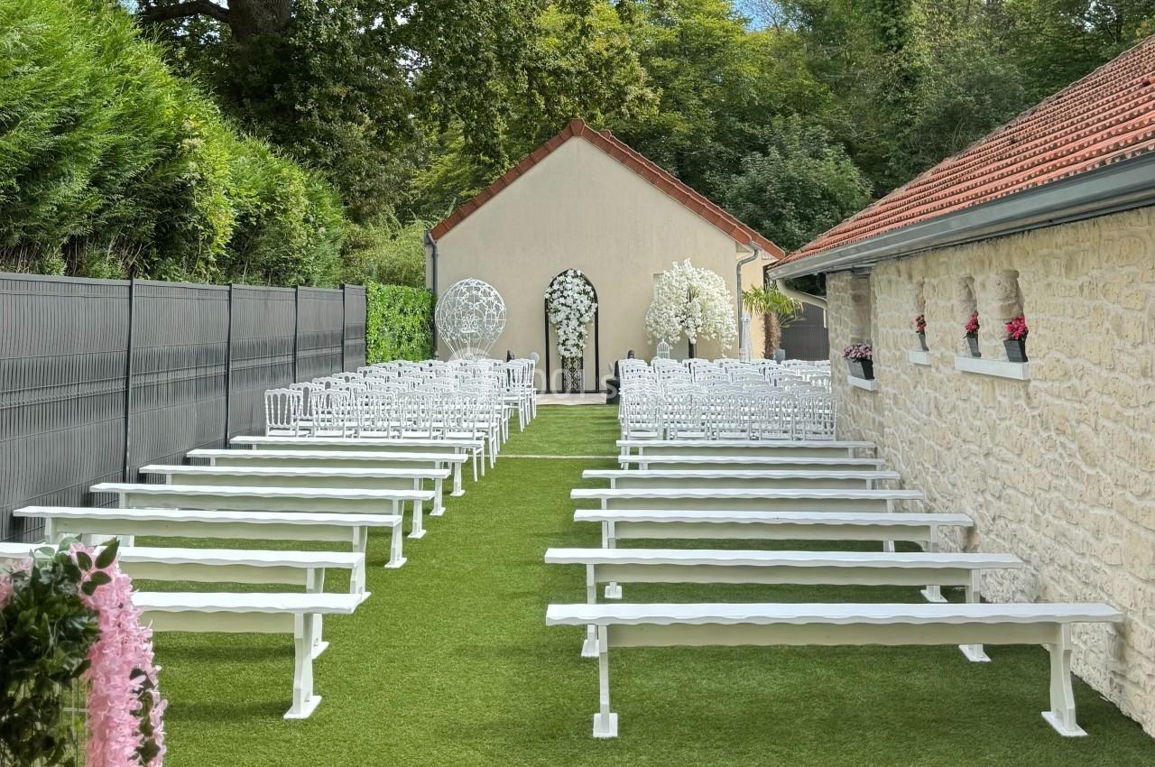 Allée centrale bordée de bancs blancs menant à une arche fleurie dans un jardin aménagé pour une cérémonie extérieure.