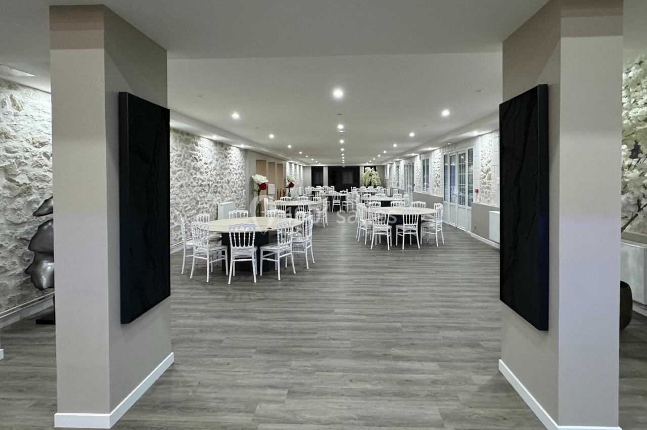 Salle de réception spacieuse avec murs en pierre, parquet, tables rondes dressées et chaises blanches.