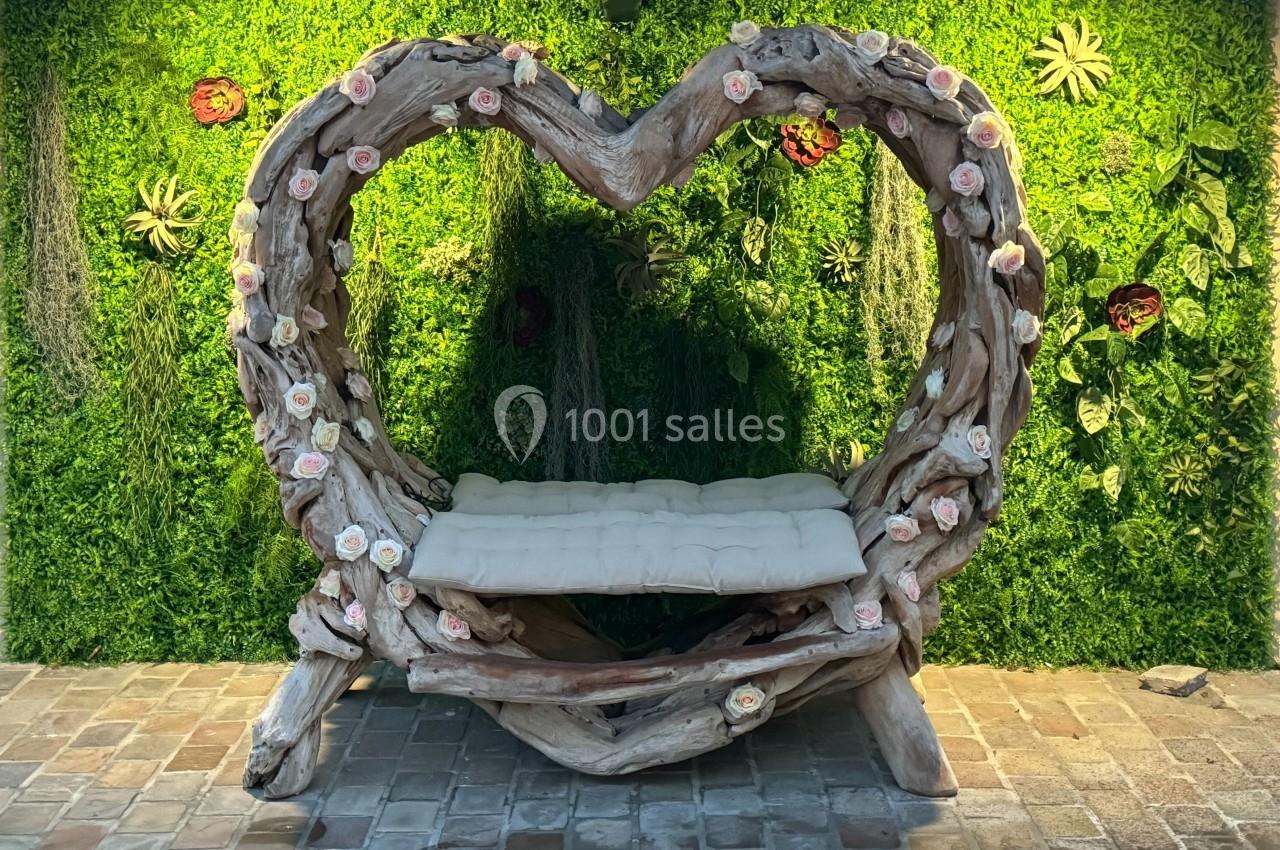Banc en bois sculpté en forme de cœur, décoré de fleurs, devant un mur végétal.