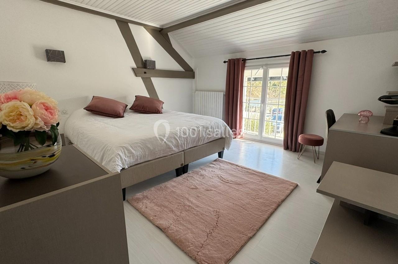 Chambre lumineuse avec lit double, bureau, rideaux roses, tapis assorti et vase de fleurs sur une commode.