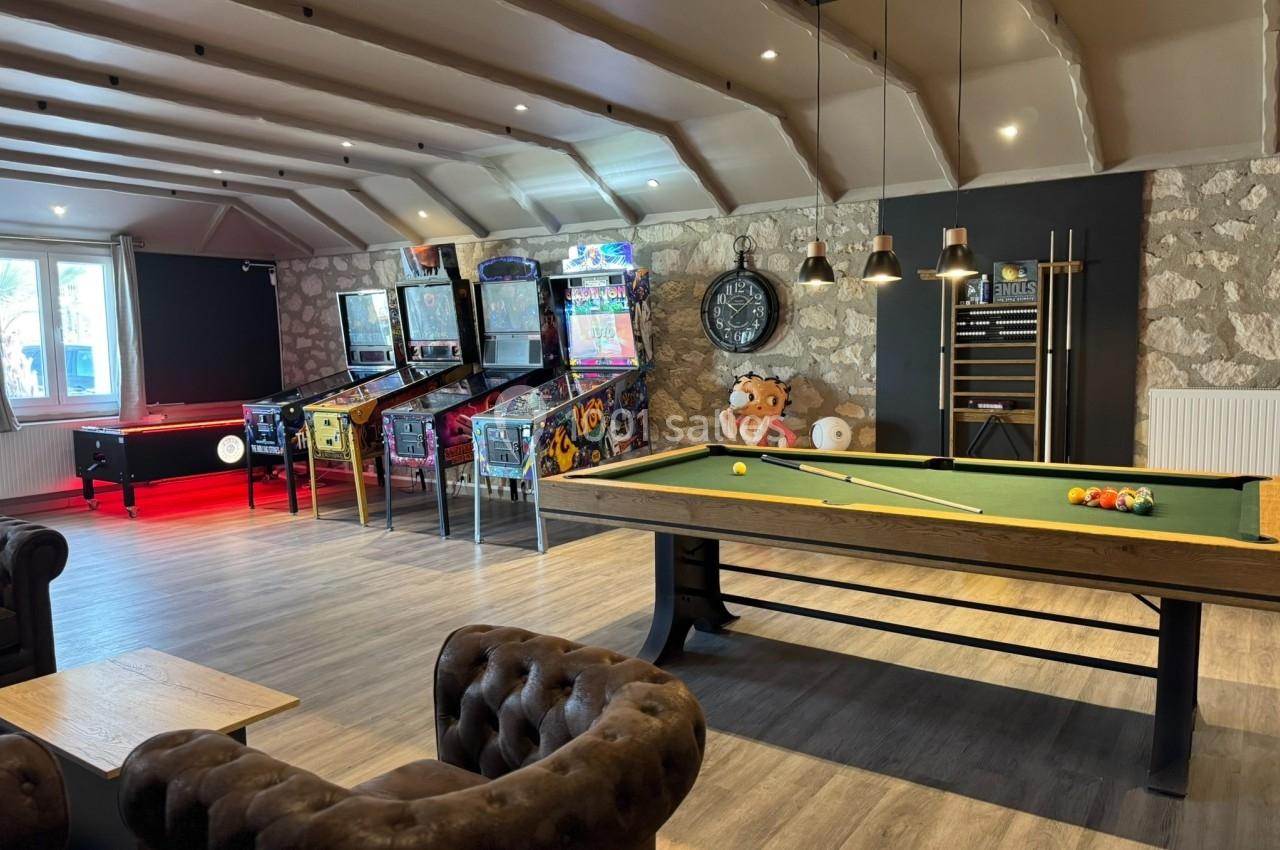 Salle de loisirs avec billard, flippers, canapé en cuir et décoration murale en pierre.