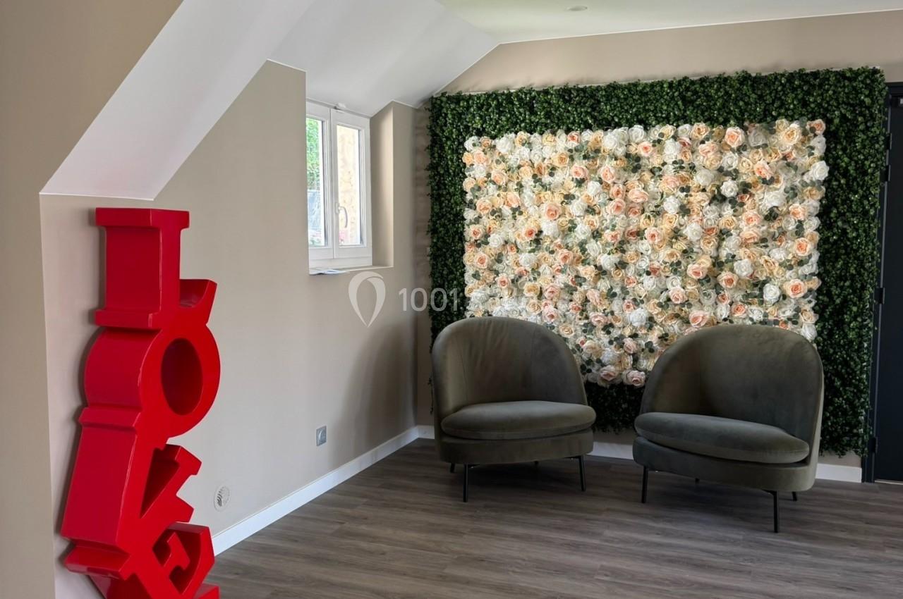 Deux fauteuils verts devant un mur végétal orné de fleurs blanches, avec une sculpture rouge ’LOVE’ à gauche.