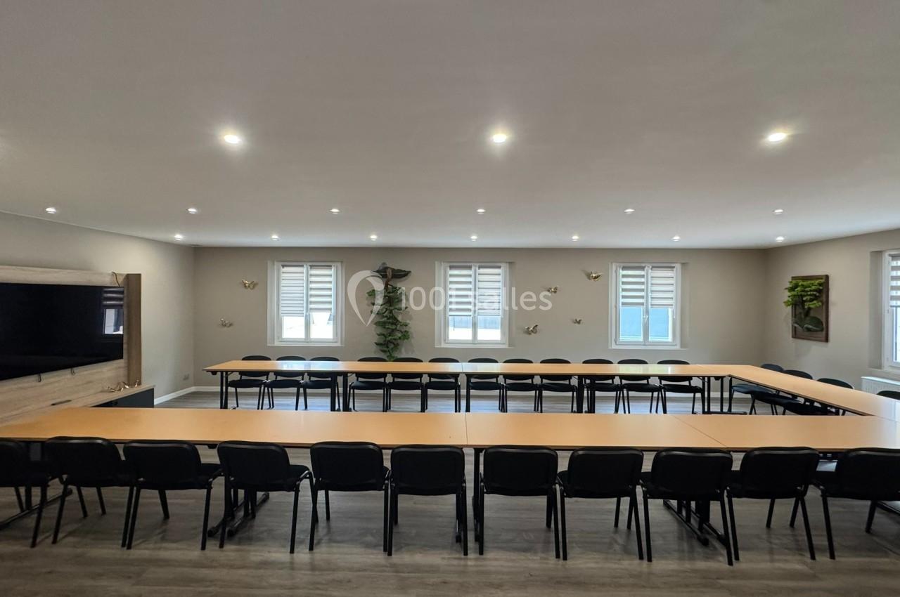 Salle de réunion lumineuse avec tables disposées en U, chaises noires, écran mural et décorations végétales.