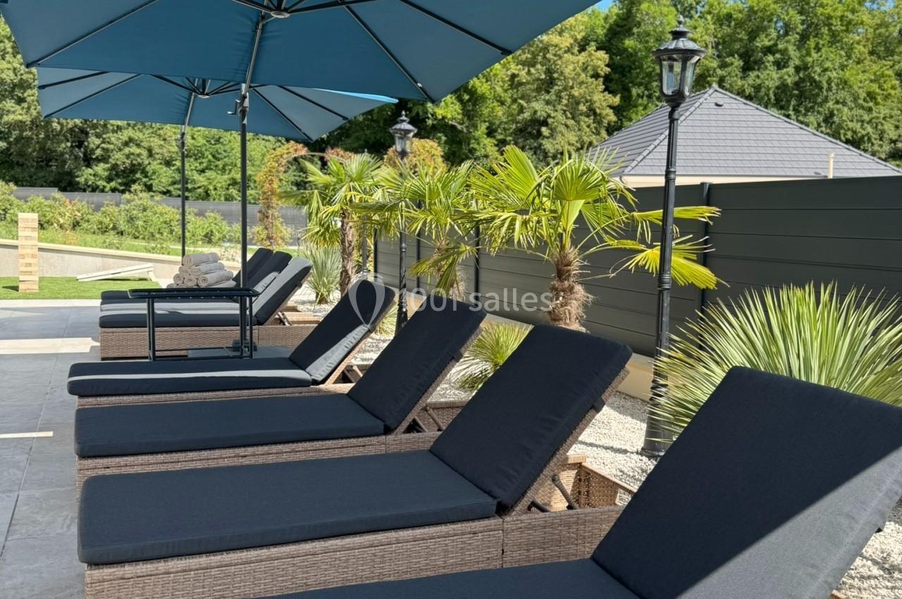 Chaises longues en rotin avec coussins noirs alignées sous des parasols bleus, entourées de végétation tropicale.
