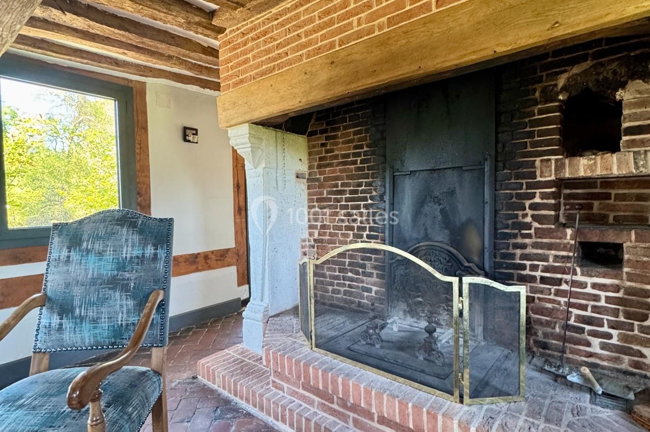 Cheminée en briques avec pare-feu métallique dans une pièce rustique, fauteuil en bois et fenêtre donnant sur l'extérieur.