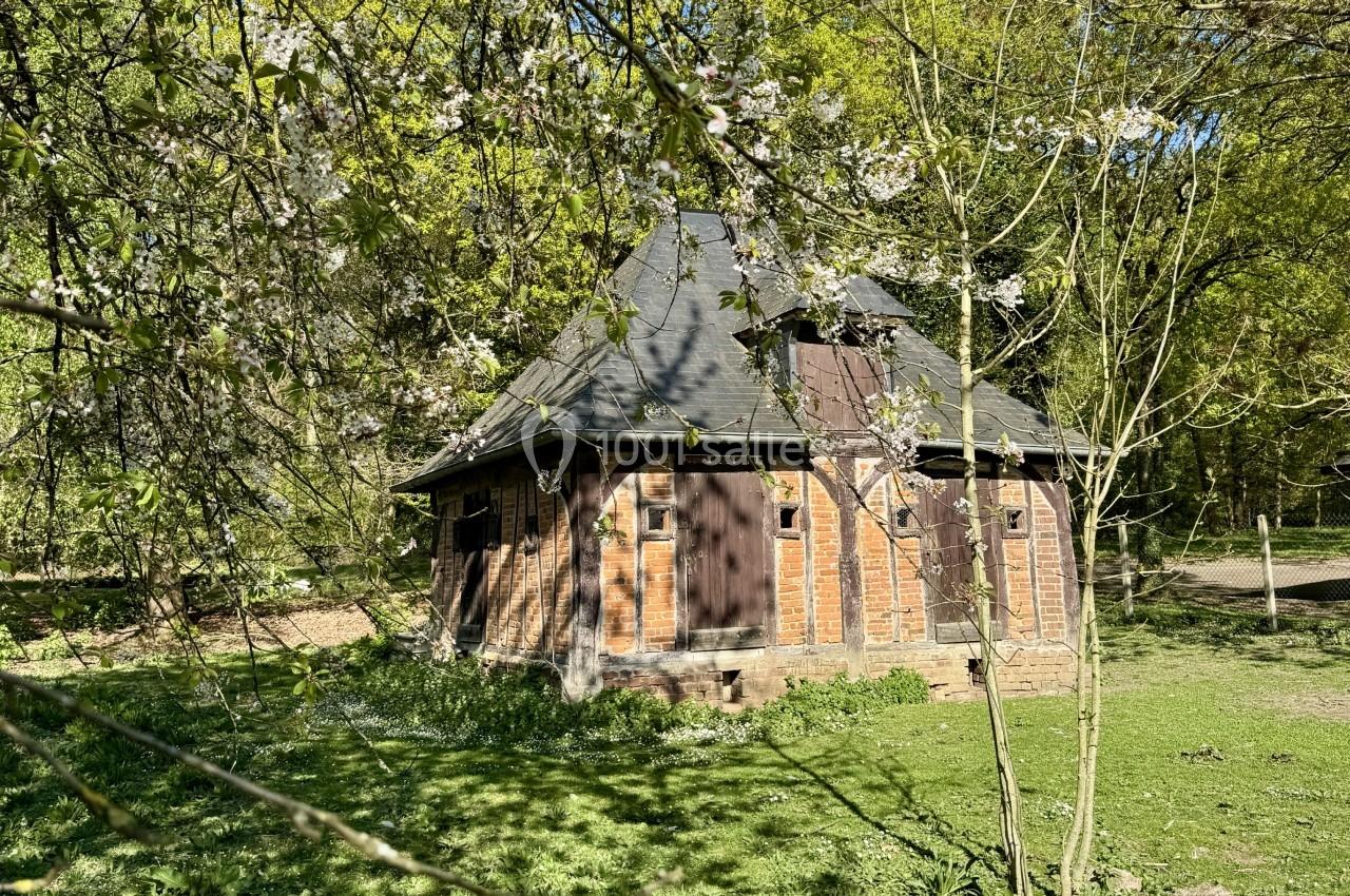 Petite maison en briques avec toit en ardoise, entourée d'arbres et de végétation sous un ciel ensoleillé.