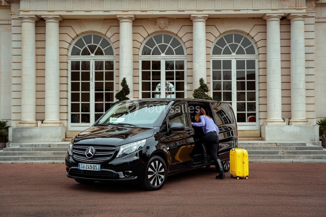 Une femme range une valise jaune dans un van noir stationné devant un bâtiment à colonnes blanches.