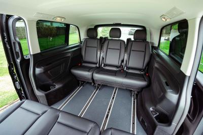 Une femme charge une valise jaune dans le coffre ouvert d'un van noir stationné dans un environnement boisé.